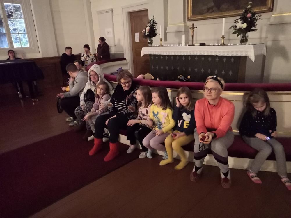 Kuvassa istuu punaisella alttarikaiteella lapsia ja aikuisia. Yö kirkossa tapahtumassa