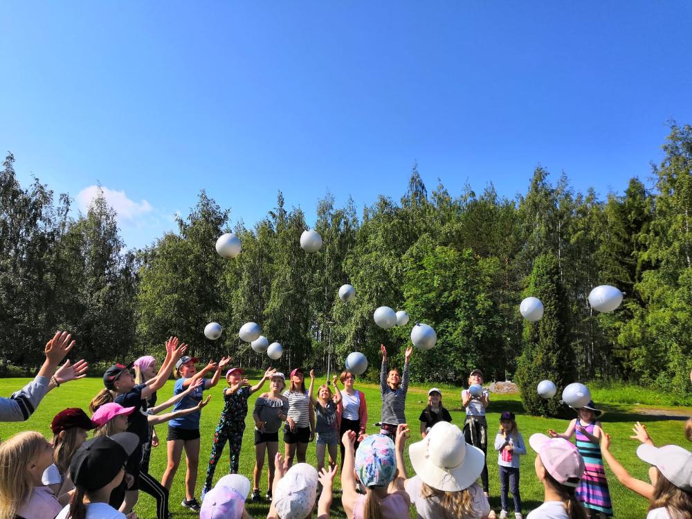 Kuvassa kesä. Lapset seisovat piirissä ja heittävät yhtä aikaa vesi-ilmapalloja ilmaan.